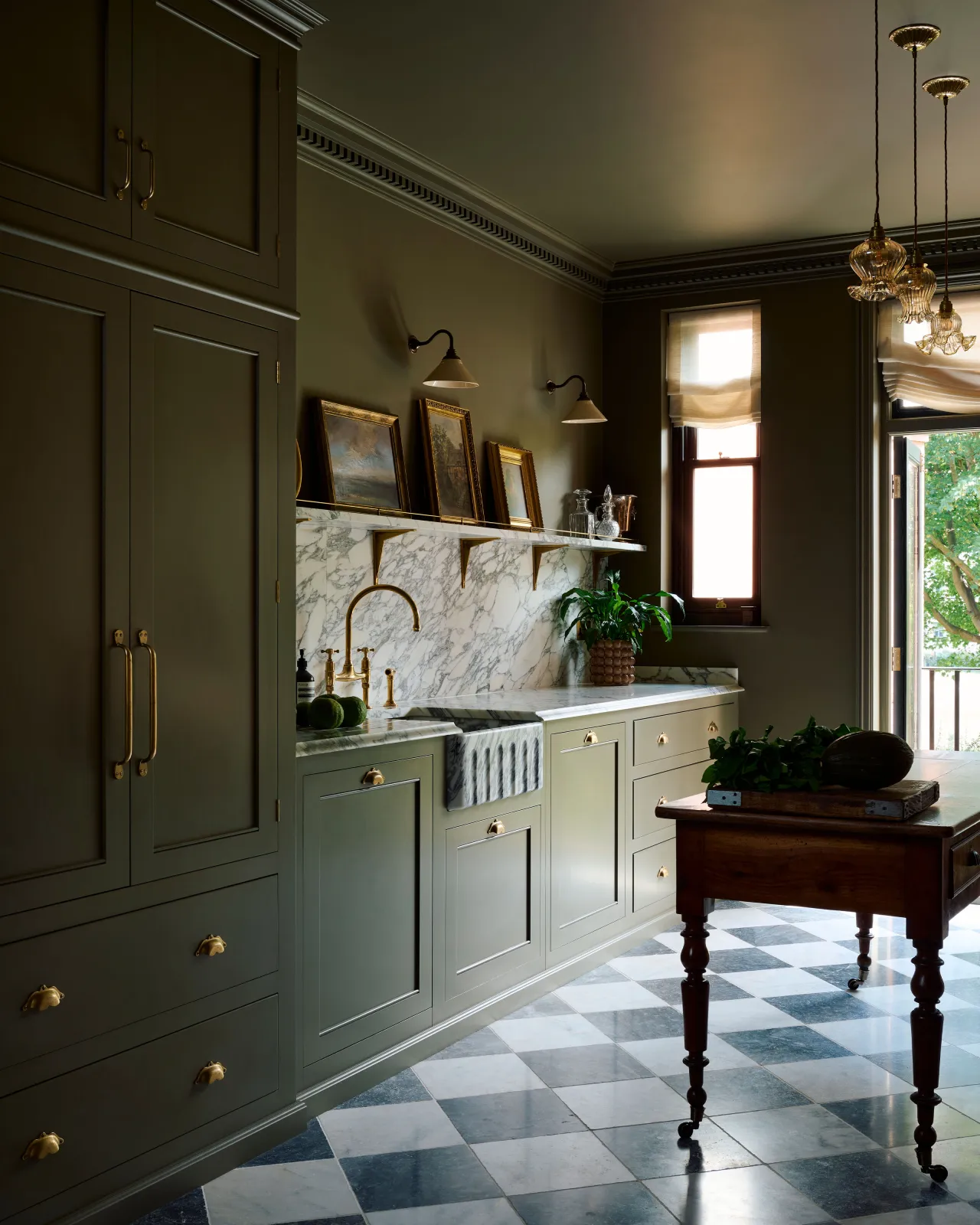 An oblique view of a classic and moody Georgian-style kitchen with mossy green walls and cabinetry, an Arabescato marble worktop, splashback and shelf and classic brass utilitarian shelf brackets.