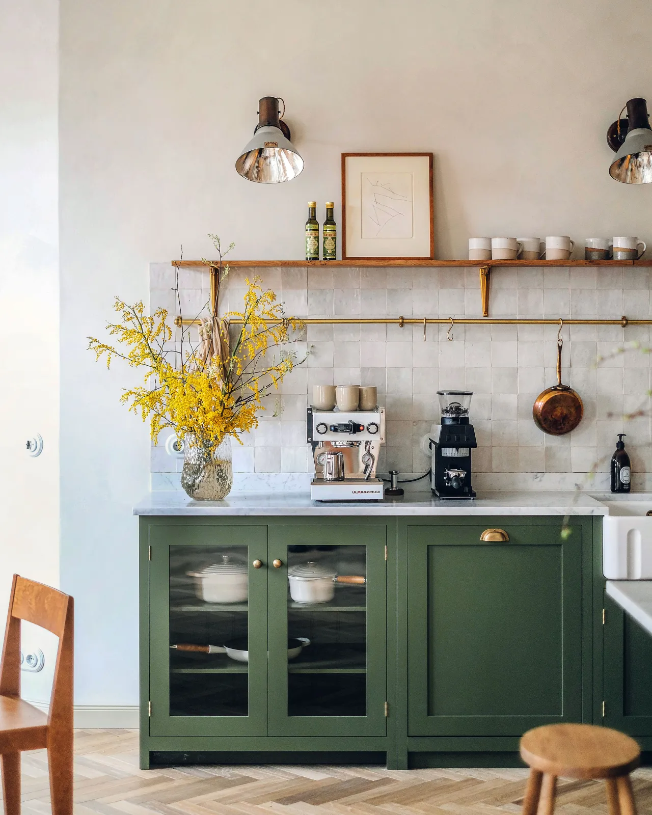 A straight-on view of a Shaker-style kitchen with racing green painted cabinetry, light plaster coloured walls, parquet wood flooring, white metro wall tiles and an oak shelf with classic brass utilitarian shelf brackets.