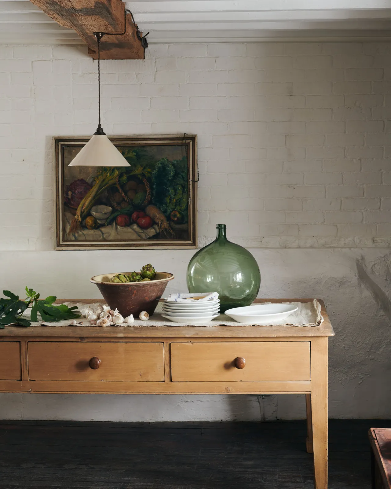 A straight-on view of a large cream glazed ceramic pendant light hanging in front of a white brick wall by an antique peach coloured prep table with drawers.