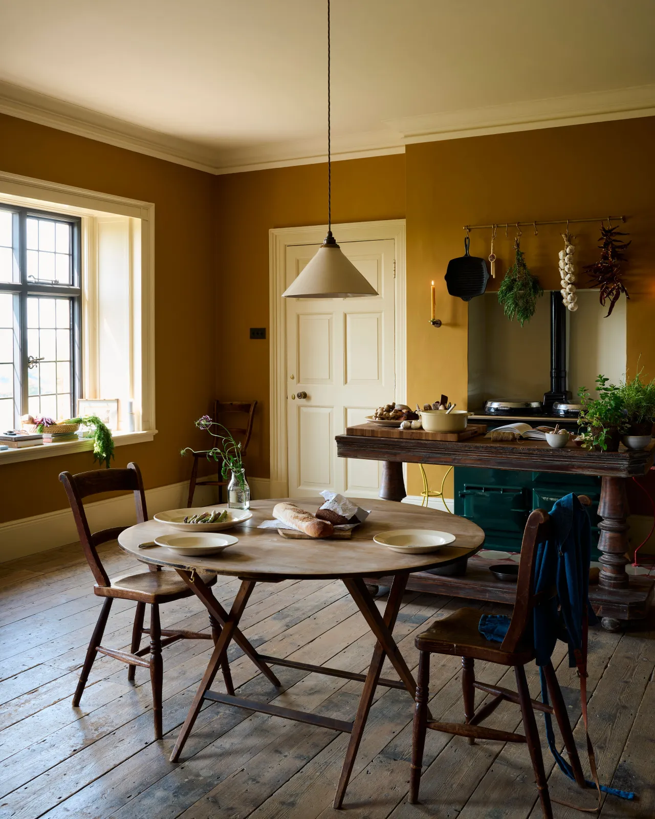 An atmospheric room with deep yellow walls, worn floorboards and a large cream glazed ceramic pendant light hanging over an antique round wooden dining table and chairs.