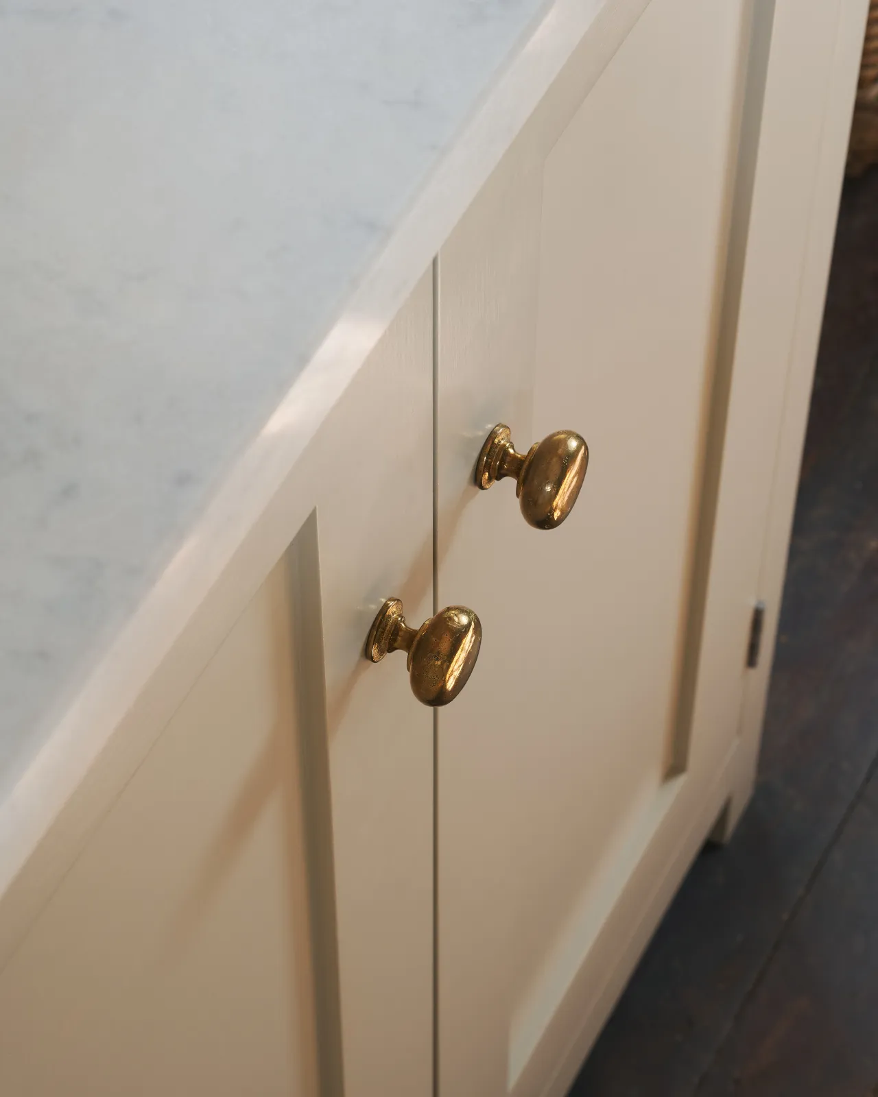 A close-up oblique view of a pair of classic knobs in a warm burnished brass finish, fitted to a mushroom painted Shaker-style kitchen cupboard.