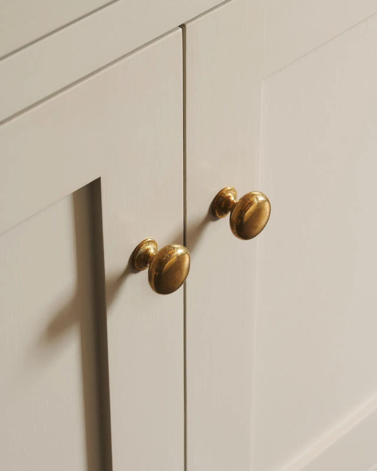 An oblique view of a pair of classic knobs in a warm burnished brass finish, fitted to a mushroom painted Shaker-style kitchen cupboard.