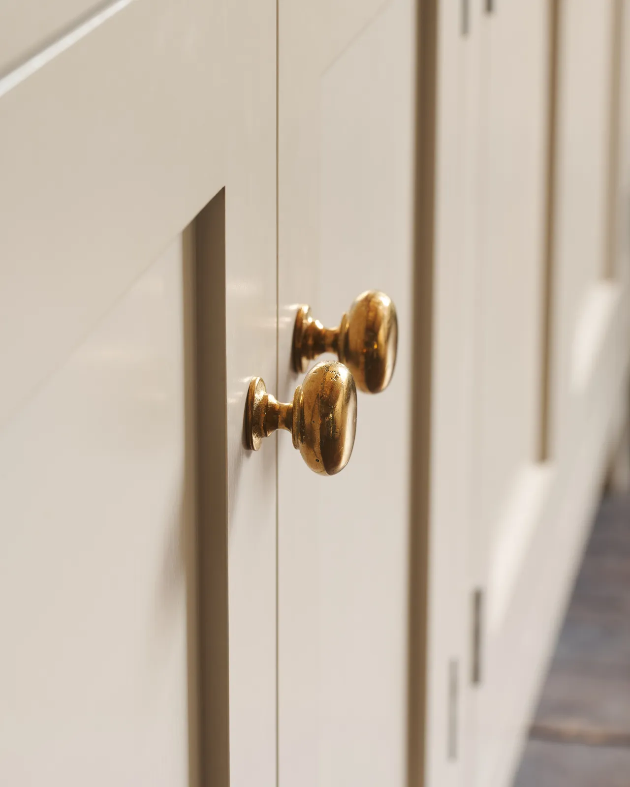 A close-up profile view of a pair of classic knobs in a warm burnished brass finish, fitted to a mushroom painted Shaker-style kitchen cupboard.