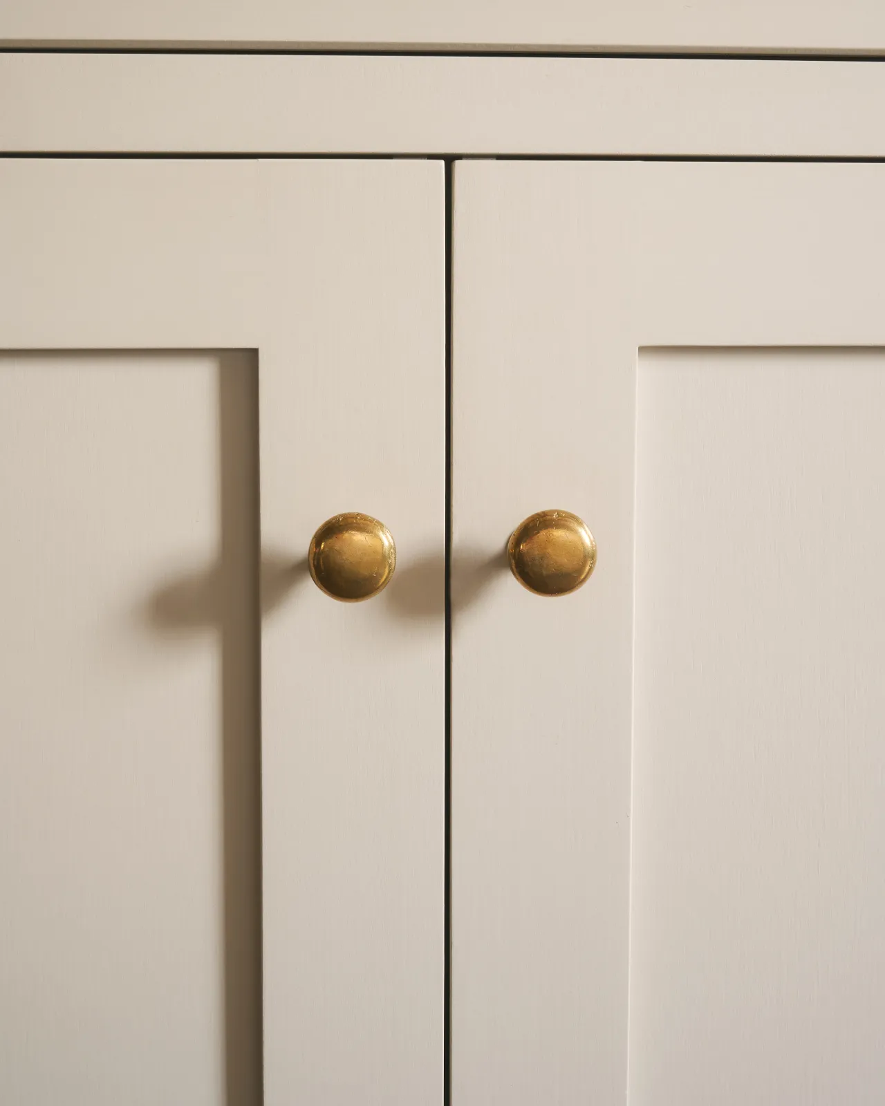 A straight-on view of a pair of classic knobs in a warm burnished brass finish, fitted to a mushroom painted Shaker-style kitchen cupboard.