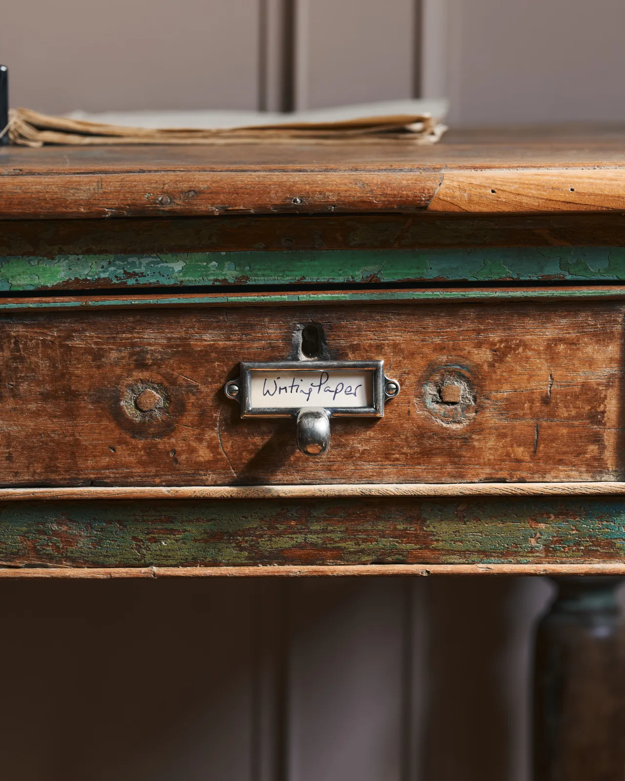 A close-up straight-on view of a small cardframe pull handle in a tarnished silver finish and a handwritten paper label that reads 'Writing Paper', fitted to the drawer of a worn antique desk table.