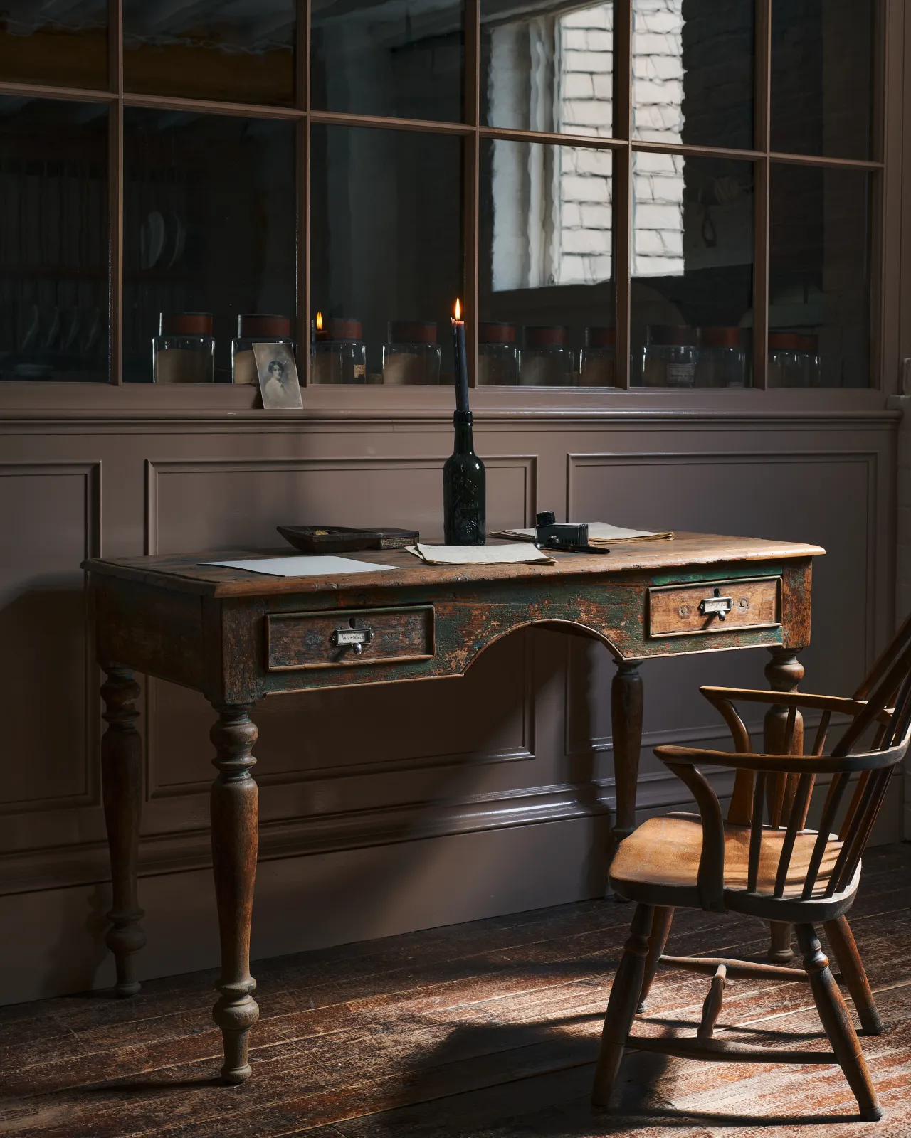 An oblique view of a dark windsor chair in front of a worn antique desk table with two small drawers, both fitted with small cardframe pull handles in a tarnished silver finish.