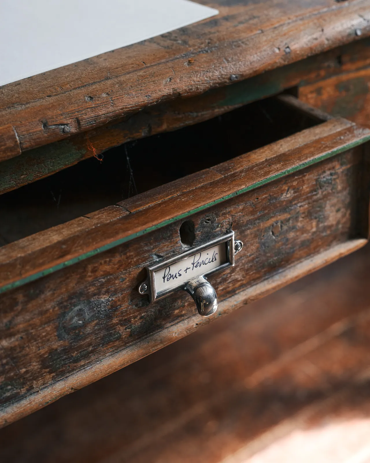 A close-up oblique view of a small cardframe pull handle in a tarnished silver finish and a handwritten paper label that reads 'Pens + Pencils', fitted to the drawer of a worn antique desk table.