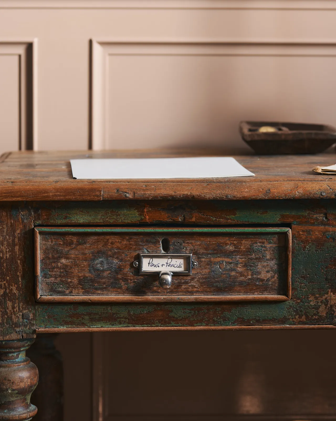 A straight-on view of a small cardframe pull handle in a tarnished silver finish and a handwritten paper label that reads 'Pens + Pencils', fitted to the drawer of a worn antique desk table.