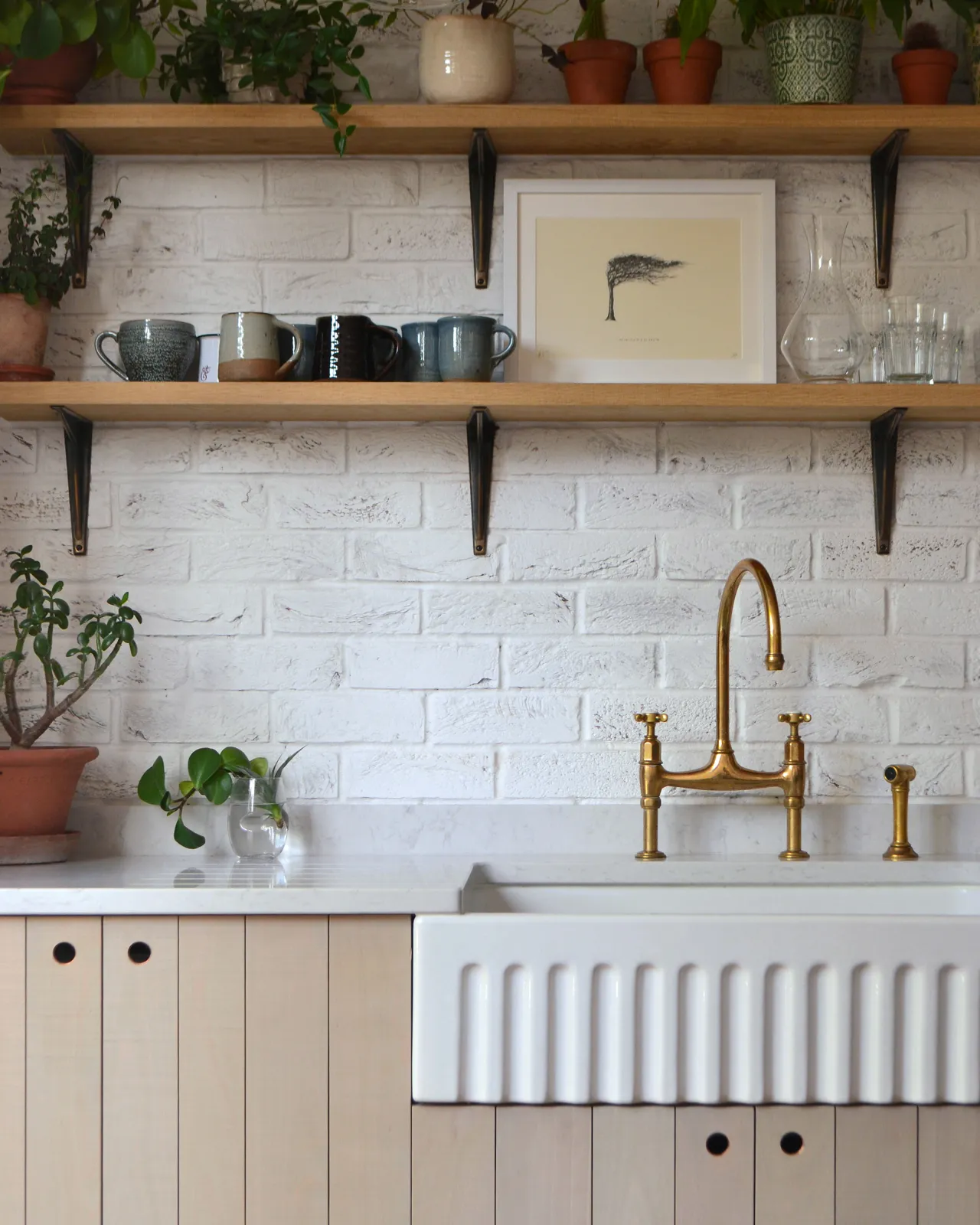 A straight-on view of a contemporary kitchen with rustic beech cabinetry, a fluted Belfast sink, white metro wall tiles and oak shelving with classic utilitarian shelf brackets in a dark oxidised brass finish.