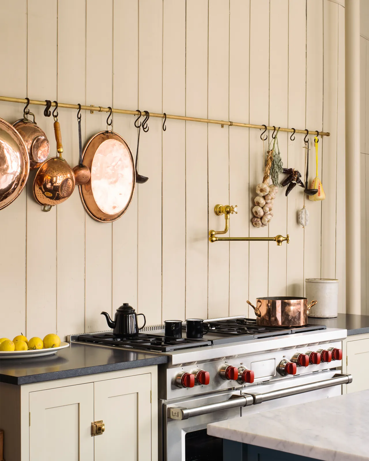 An oblique view of a large stainless steel range cooker below a long aged brass hanging rail with a large collection of vintage copper pans and utensils hanging from black oxidised brass s hooks.