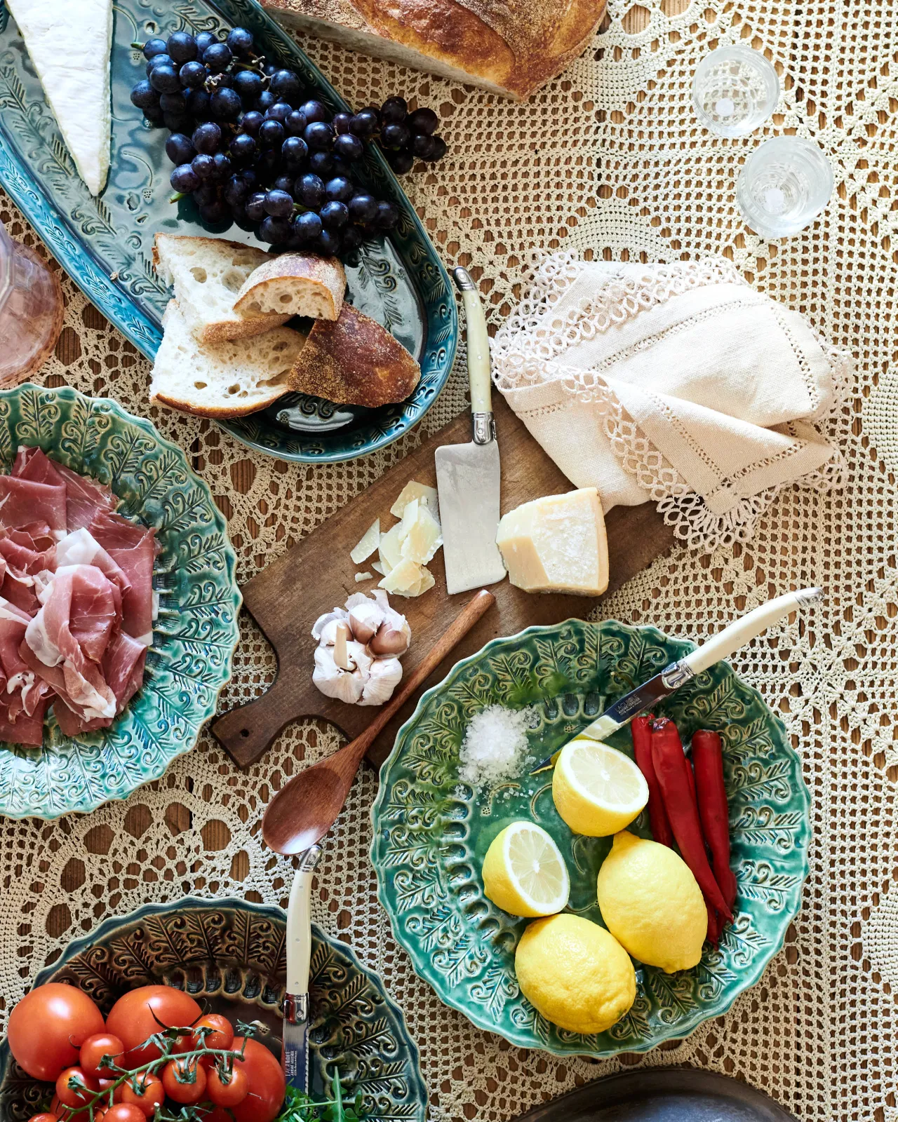 An overhead view of several oval and round handmade bohemian-style ceramic platters, with black grapes, cheeses, homemade bread, lemons, chillis and prosciutto ham on a beautiful crochet lace tablecloth.