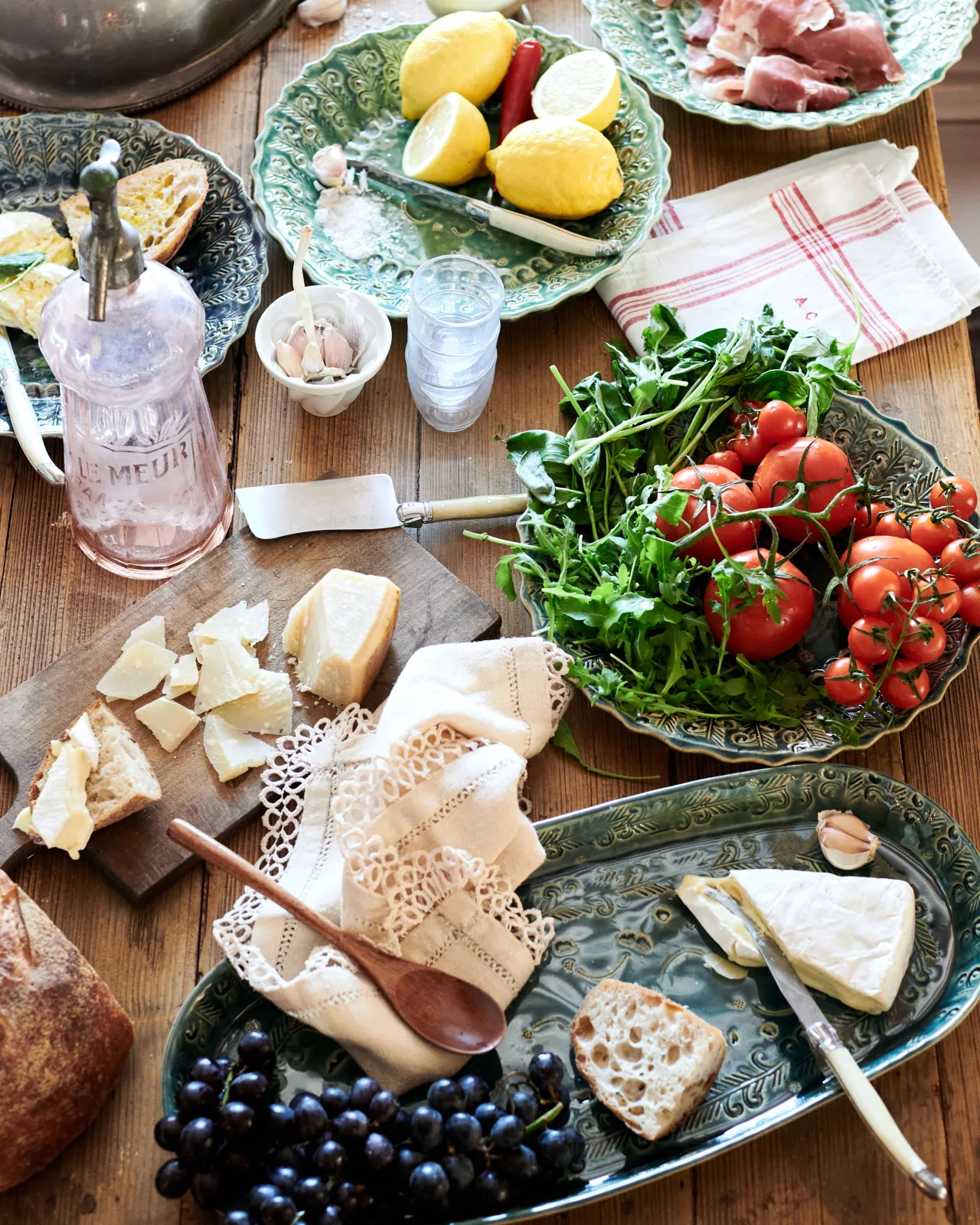 Several oval and round handmade bohemian-style ceramic platters, with black grapes, cheeses, homemade bread, lemons, chillis, prosciutto ham, tomatoes and leafy greens.