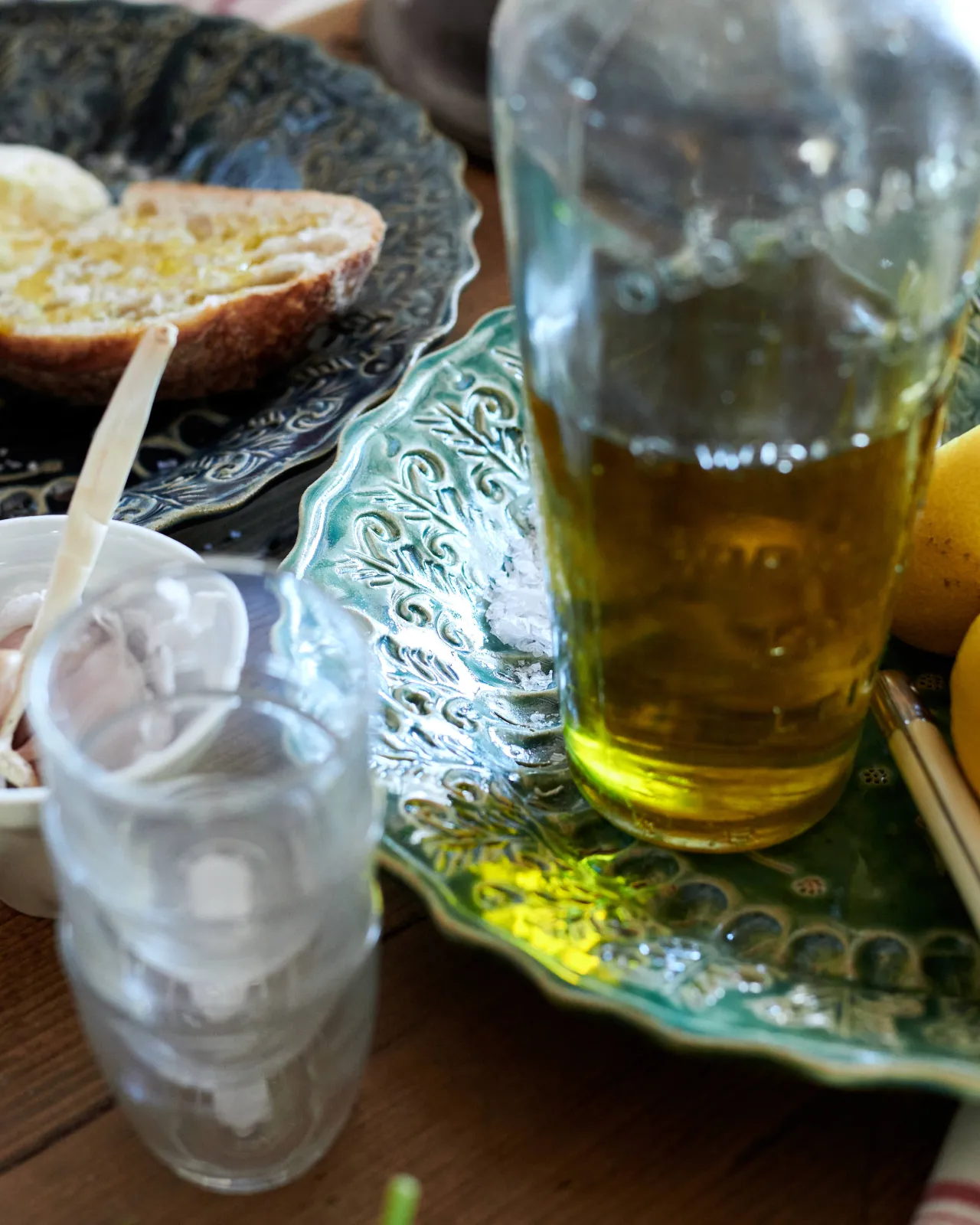 A close-up view of a clear bottle of oil on a round handmade bohemian-style green ceramic platter with delicate impressions and surface patterns.