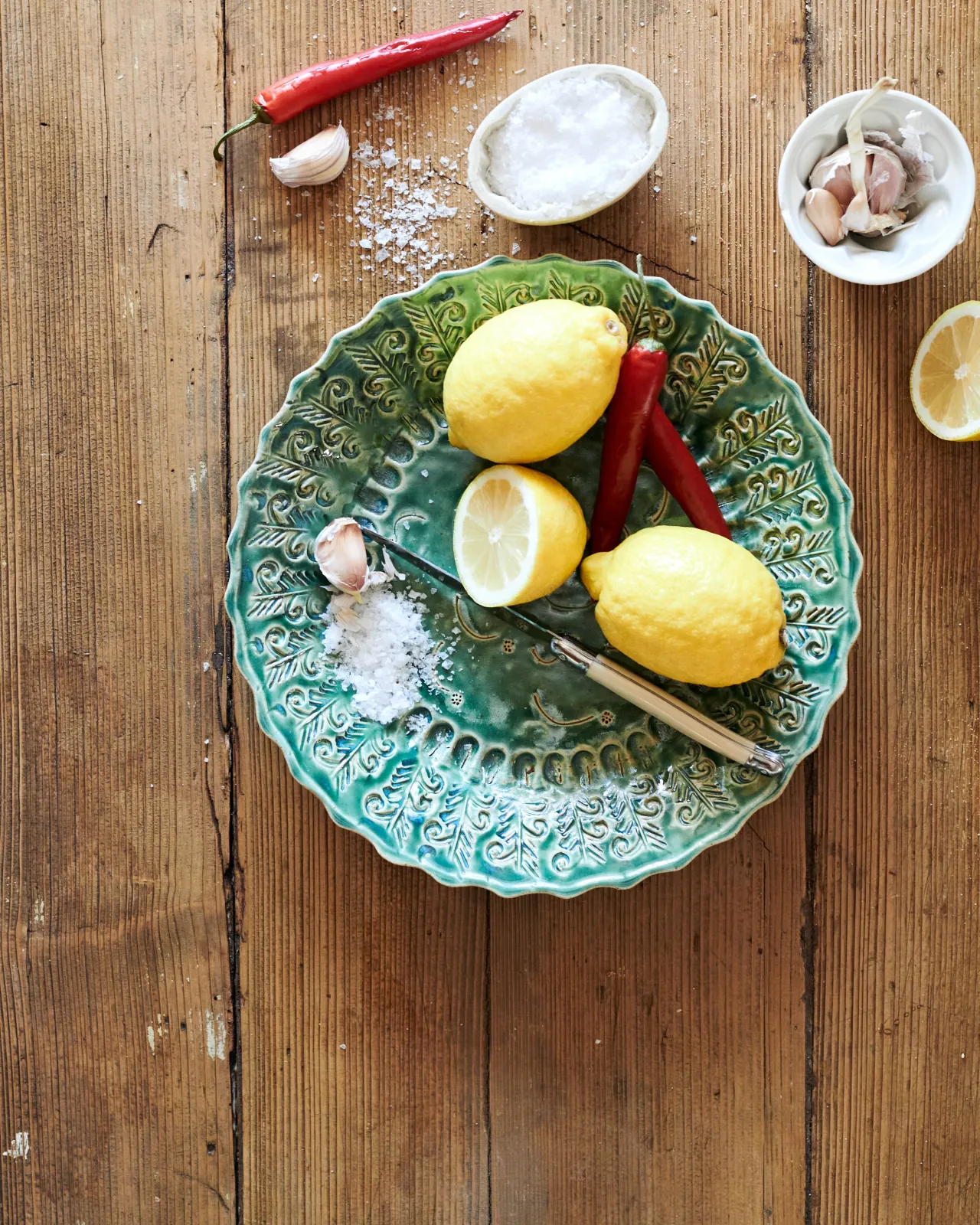 An overhead view of a round handmade bohemian-style green ceramic platter with delicate impressions and surface patterns on a rustic pine tabletop with vintage silverware, lemons, salt, chillis and garlic.