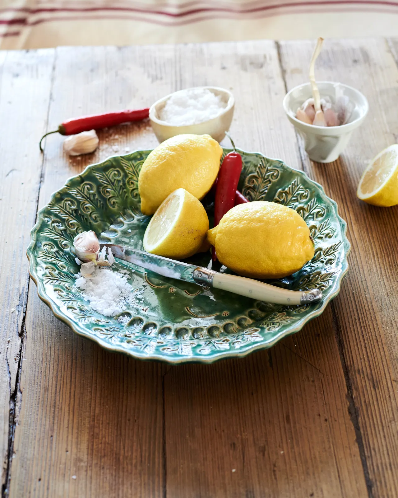 A round handmade bohemian-style green ceramic platter on a rustic pine tabletop with vintage silverware, lemons, salt, chillis and garlic.