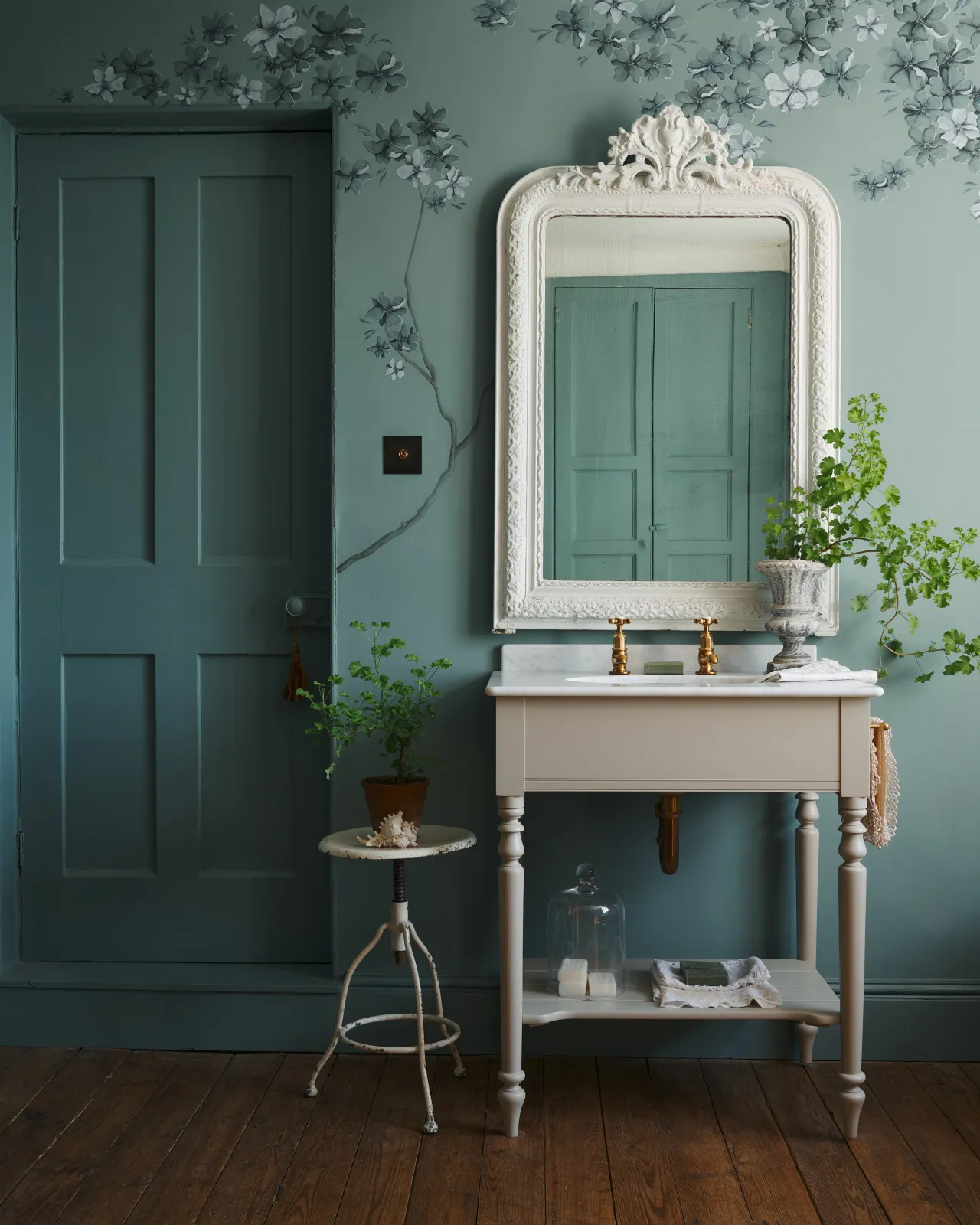 A straight-on view of a mushroom-grey Victorian-inspired washstand with brass pillar taps in a duck-egg blue painted room. The room is classic and bohemian with an ornate white mirror and elegant painted flowers across the top of the walls.