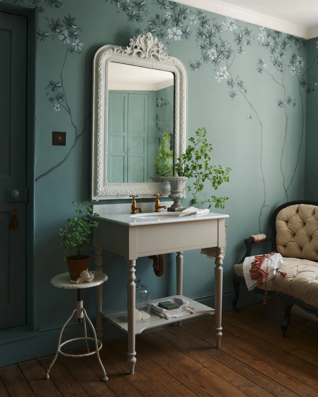 An oblique view of a mushroom-grey Victorian-inspired washstand with brass pillar taps in a duck-egg blue painted room. The room is classic and bohemian with an ornate white mirror and elegant painted flowers across the top of the walls.
