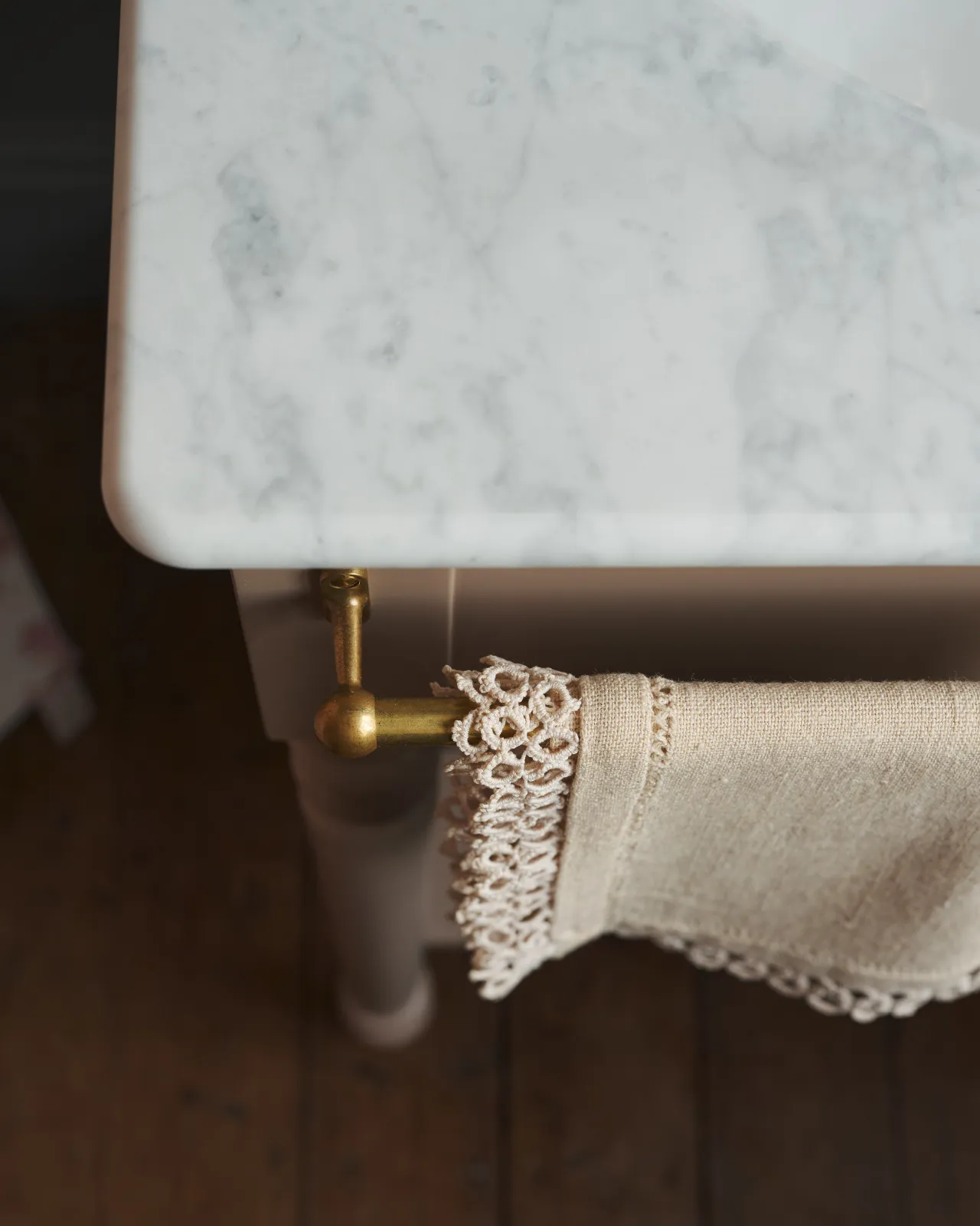 A detailed view of an aged brass towel rail attached to the front of a Victorian-inspired washstand, a vintage lace hand towel hangs from the rail.