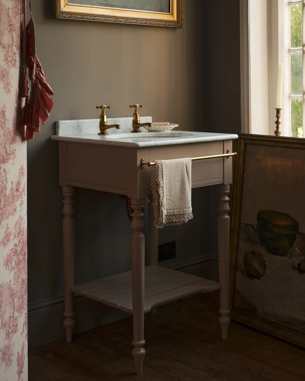 An oblique view of a dusty-pink Victorian-inspired washstand with brass pillar taps in a greyish-brown painted room. The shot is atmospheric and moody, a vintage lace towel hangs from a brass rail attached to the front of the washstand.