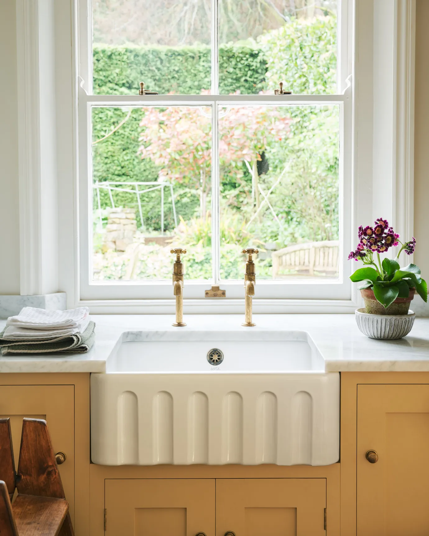 A close-up straight-on view of a small fluted ceramic Belfast sink in a yellow painted Shaker-style kitchen with aged brass bibcock taps.