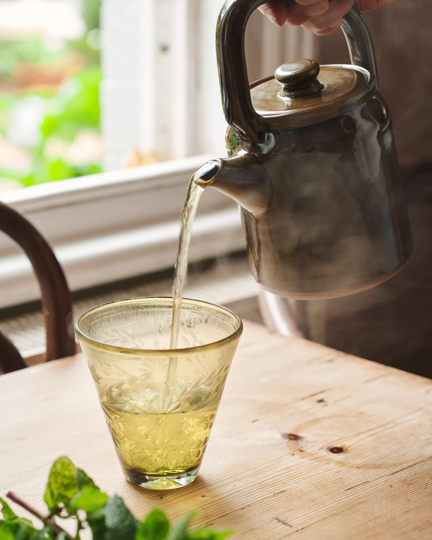 A small mid-century-style ceramic teapot with a glossy amber glaze pouring tea into a conical yellow glass.