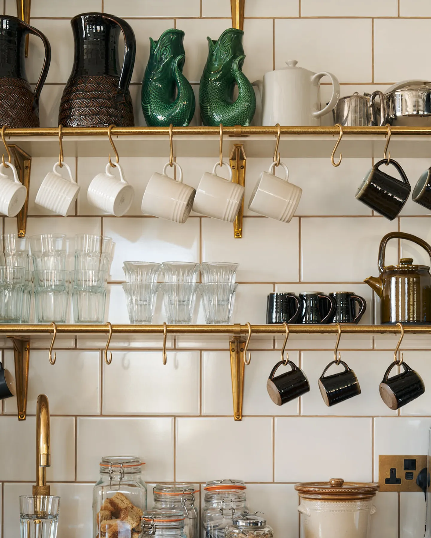A close-up shot of rows of shelves fitted to the back of a little drinks-making station. Aged brass rails are attached to the shelves and ‘s’ hooks provide the perfect place to hang coffee cups. The image is beautifully vibrant with glowing brass, glossy ceramics and reflective glassware.