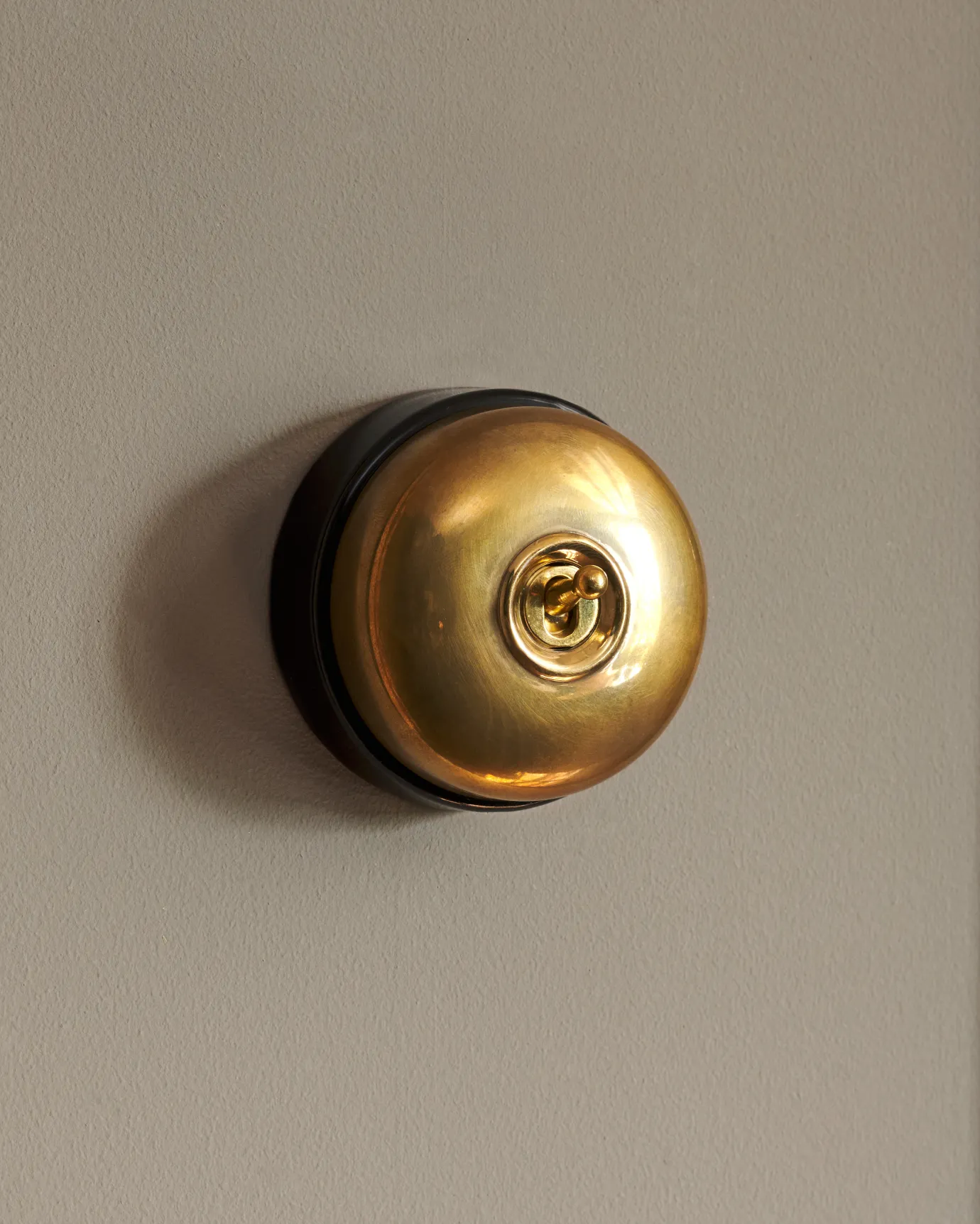 A side-on view of a small 1 gang round-shaped toggle switch in a warm shiny brass finish with a black base.