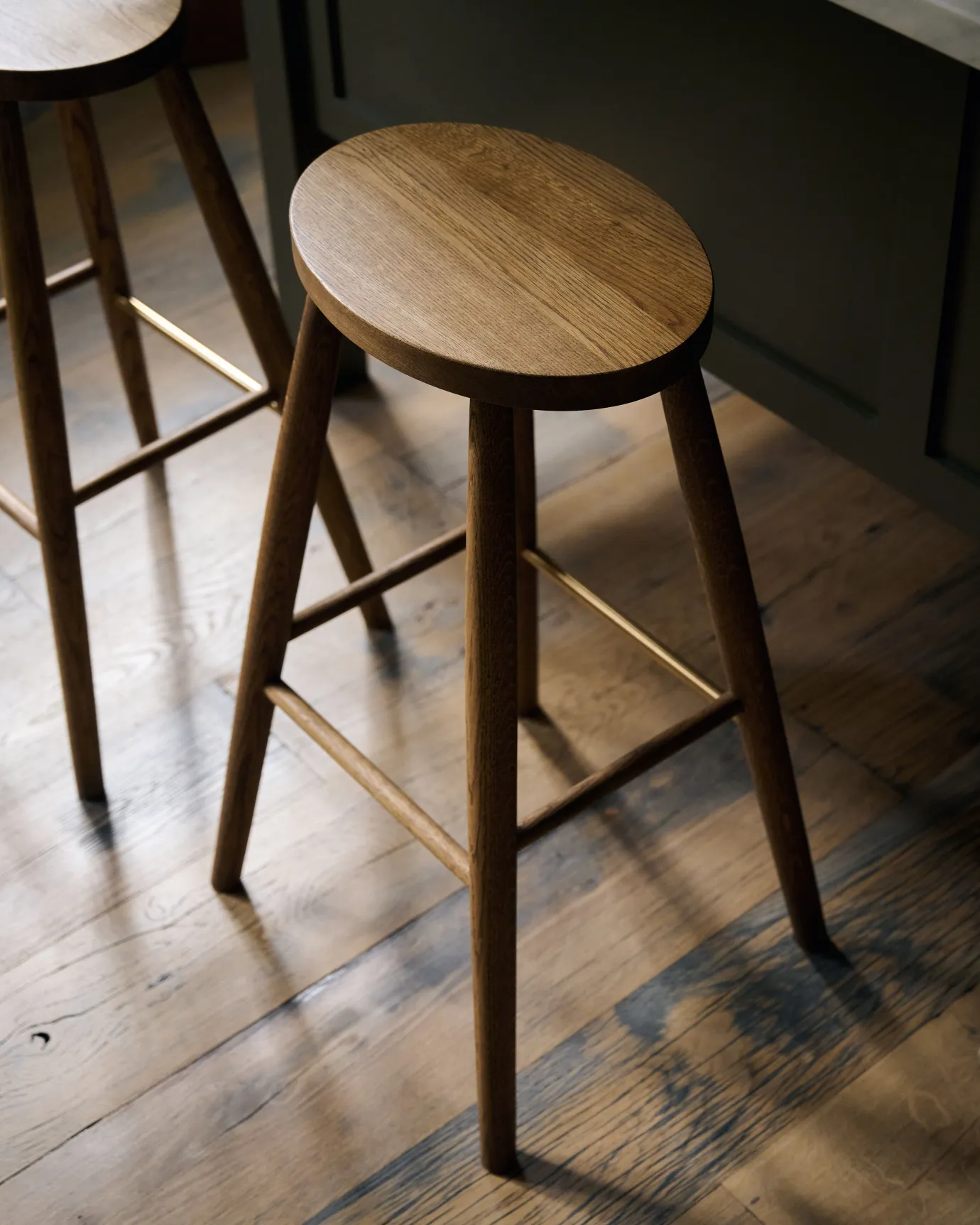 An oblique view of a simple oak bar stool with an oval-shaped seat and a brass footrail.