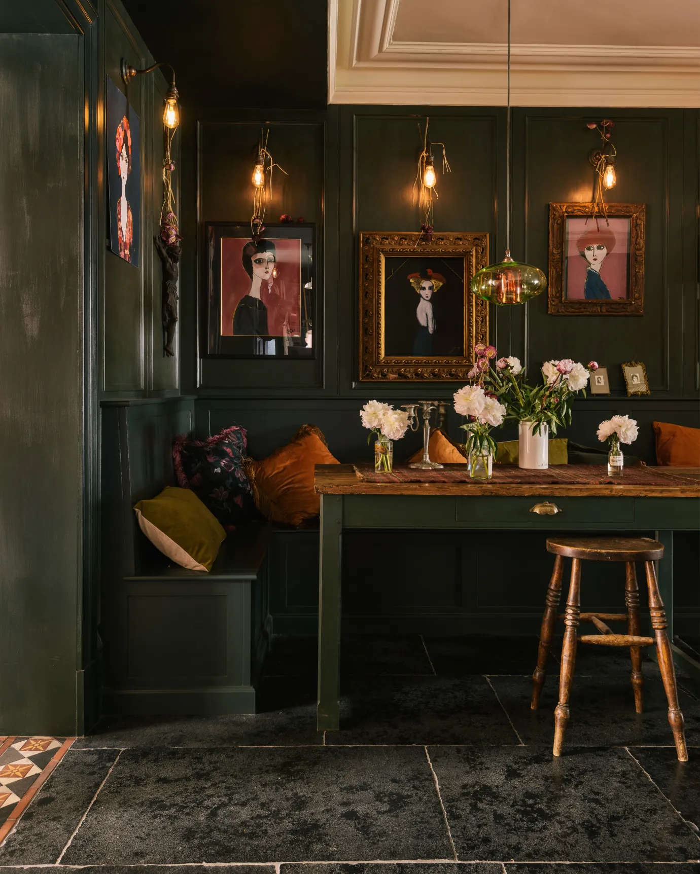 A view towards a moody and atmospheric dining area, with built-in wooden bench seating and antique-style Shaker table, all painted in dark green. Light trickles in from an open door, highlighting the worn texture and colour variation on the Brushed Charcoal Limestone tiles.