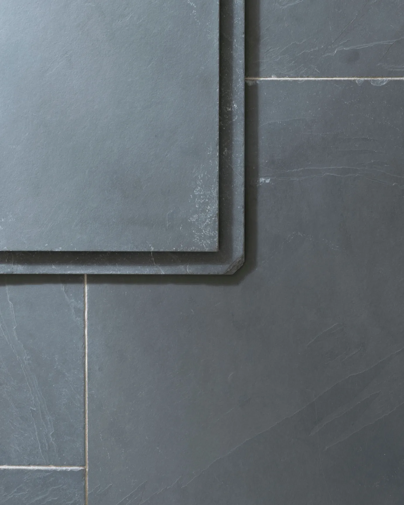 A close-up overhead view of a grey tumbled slate floor, with two loose tiles stacked in one corner.