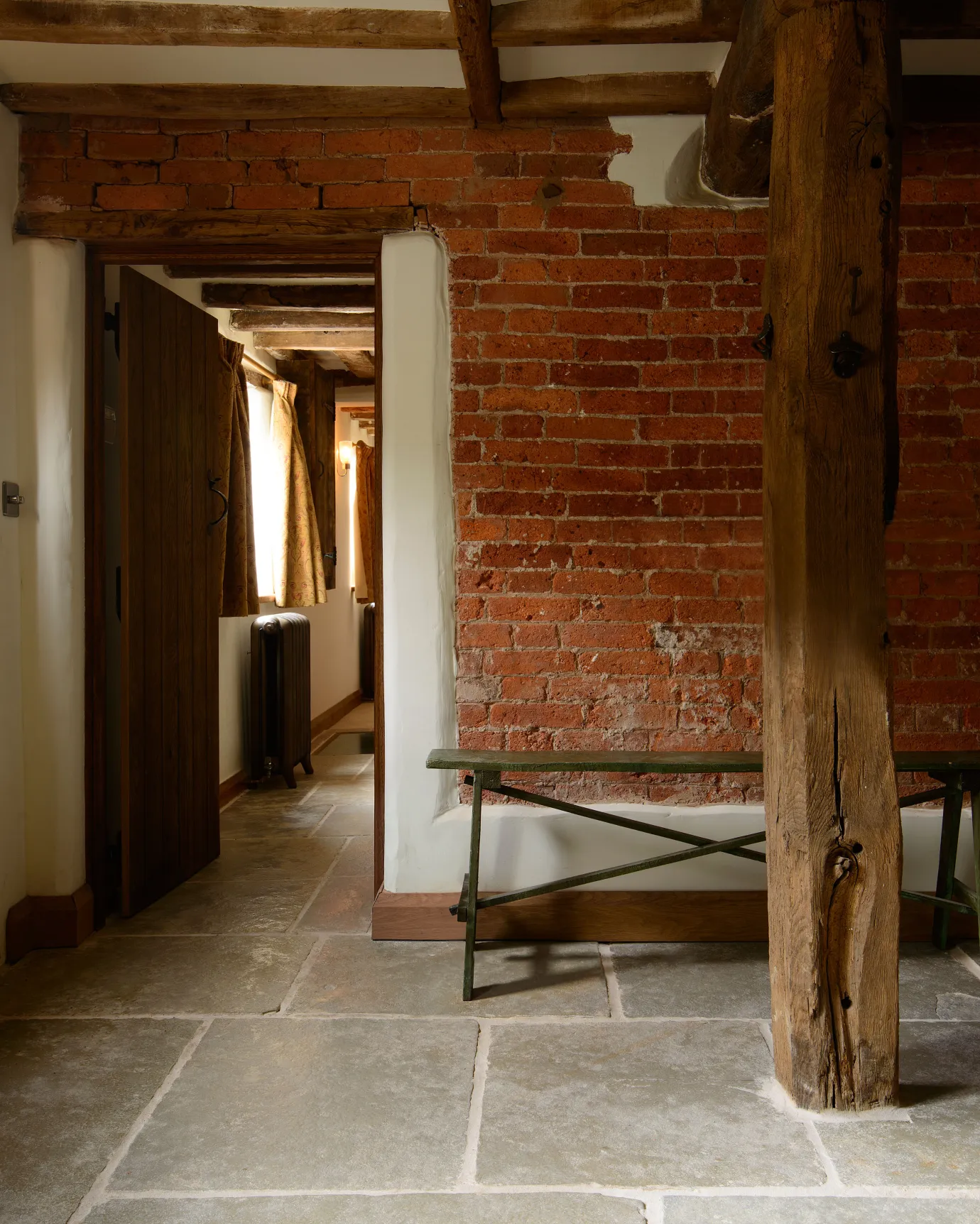 A cottage entranceway with red brick walls, wooden beams and an aged limestone floor with natural earthy tones of green, brown and grey.